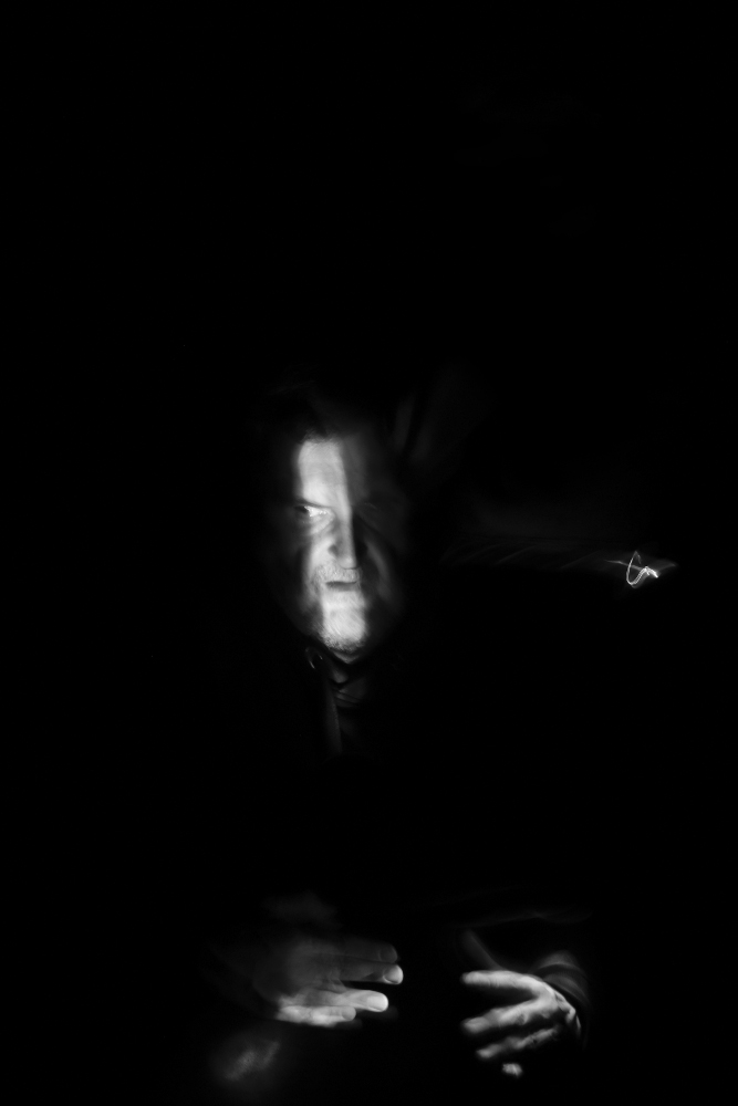 An-Sich-Halten (Selbstportrait) 2020 von Gerald Pirner Das schwarzweiße Light Painting wurde im Hochformat aufgenommen und zeigt als zentrales Motiv einen Mann im Dunkeln, der von einem schmalen Lichtstreifen bestrahlt wird. Sein Kopf befindet sich im Mittelpunkt des Bildes und ist leicht unscharf. Nur die linke Gesichtshälfte und sein Nasenrücken werden teilweise von einem länglichen hellen Lichtstrahl beleuchtet. Der Mann blickt mit seiner linken Pupille nach links ins Dunkel. Er trägt einen Vollbart und sein geschlossener Mund wirkt leicht zusammengekniffen. Am unteren Bildbereich fällt ein sanfter Lichtstrahl auf zwei Hände, deren Handrücken dem Betrachter zugewandt sind. Die Finger beider Hände sind in einer verkrampften Position, was als Festhalten eines unsichtbaren Gegenstandes gedeutet werden kann. Arianna Waldner Bingemer