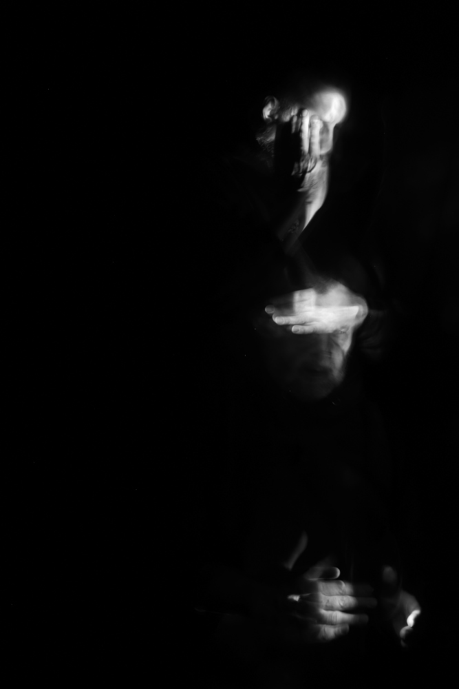 Der Turm der Hände (Doppelselbstportrait) 2020 von Gerald Pirner Die Schwarz-weiß-Fotografie im Hochformat wurde mithilfe der Lightpainting-Technik hergestellt. Oben rechts im Bild sieht man ein Gesicht im Dreiviertelprofil, wobei Auge und Mund von einer Hand verdeckt werden. Darunter befindet sich ein weiteres Gesicht, bei dem die Augen von einer hell ausgeleuchteten Hand verdeckt werden. Unten rechts im Bild sieht man zwei zueinander zeigende Hände. Der Hintergrund ist schwarz.
